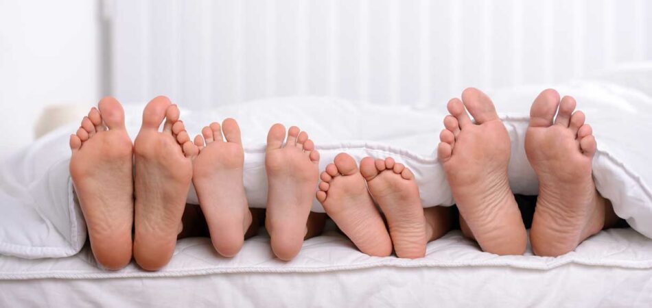 Mother, Father And Two Children Lie On Bed With White Sheets; Focus On Feet